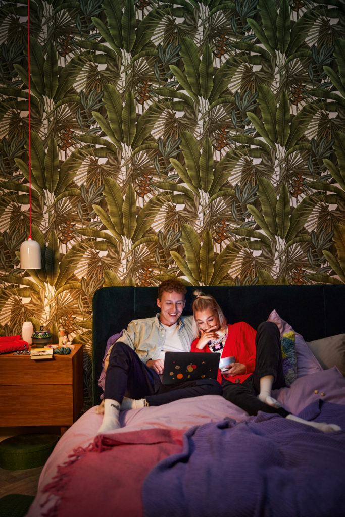 Cette image présente deux personnes regardant un ordinateur en mangeant dans une chambre. Le papier peint illustre des grandes palmes et feuillages tout droit sortis de la jungle.
